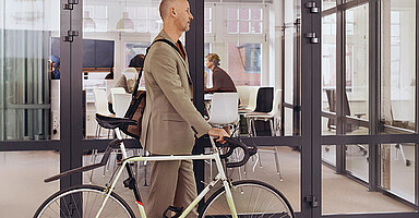 Grenzmanagement: Mann im Anzug schiebt ein Fahrrad durch ein modernes Büro mit Glaswänden. Grenzmanagement: Mann im Anzug schiebt ein Fahrrad durch ein modernes Büro mit Glaswänden.