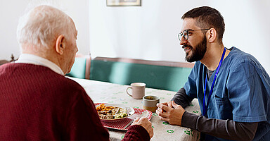 Ausländische Fachkräfte: Ein Pfleger sitzt bei einem Senior, der gerade isst Ausländische Fachkräfte: Ein Pfleger sitzt bei einem Senior, der gerade isst