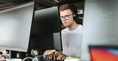 Neurodiversität: Blick auf einen jungen Mann durch Monitore hindurch, der hochkonzentriert ist und Kopfhörer trägt. Neurodiversität: Blick auf einen jungen Mann durch Monitore hindurch, der hochkonzentriert ist und Kopfhörer trägt.