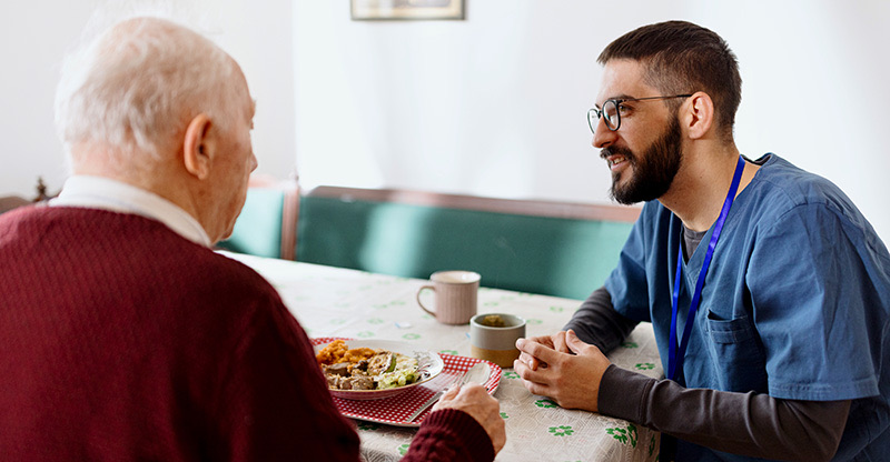 Ausländische Fachkräfte: Ein Pfleger sitzt bei einem Senior, der gerade isst Ausländische Fachkräfte: Ein Pfleger sitzt bei einem Senior, der gerade isst