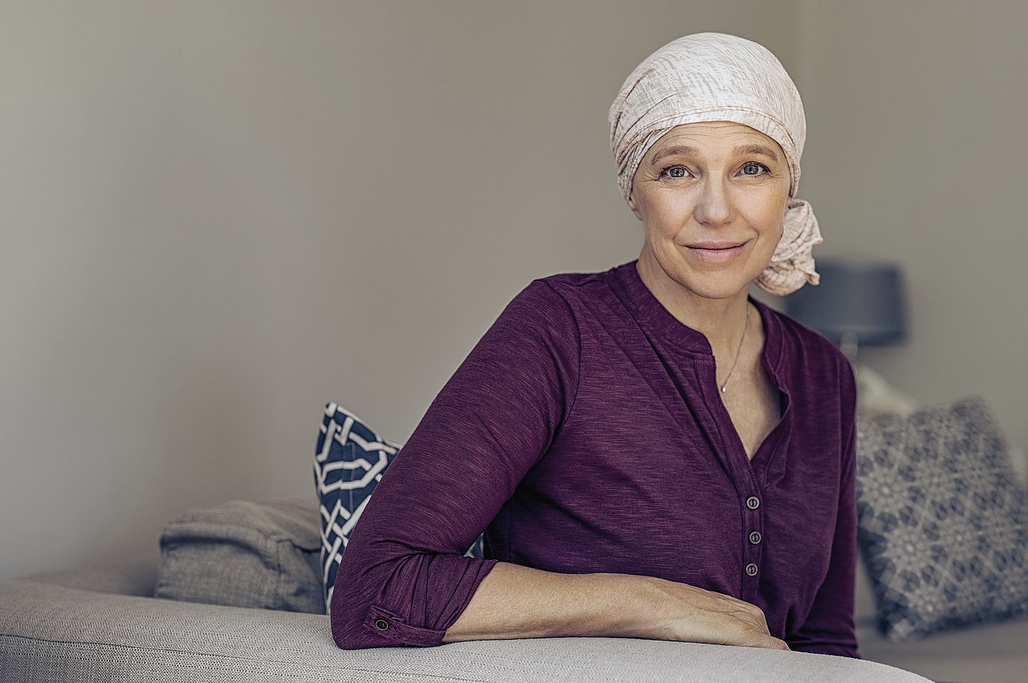Foto einer Frau auf dem Sofa sitzend, die sich an der Rückenlehne abstützt, mit einem Kopftuch, das sie offensichtlich nach einer Chemotherapie tragen muss