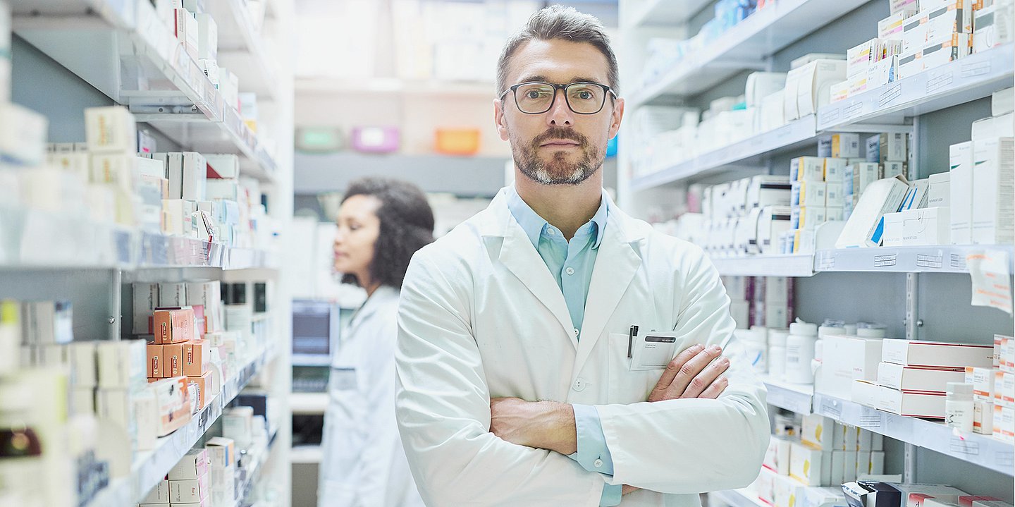 Foto: Ein Mann in weißem Kittel steht zwischen Regalen, die mit Medikamenten gefüllt sind.