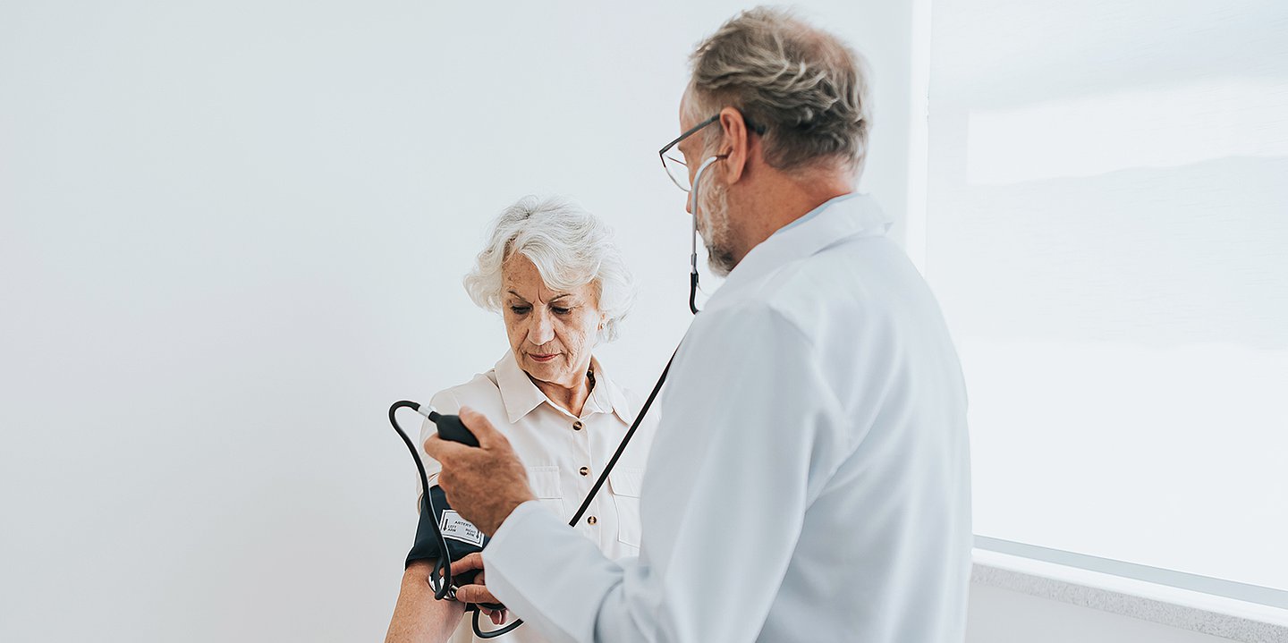 Foto: Ein Mann im weißen Kittel (Arzt) misst einer älteren Frau den Bluttdruck.