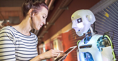 KI und Arbeit: Frau mit Tablet vor einem humanoiden Roboter KI und Arbeit: Frau mit Tablet vor einem humanoiden Roboter