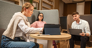 Zwei Frauen und ein Mann sitzen an einem Tisch und schauen auf einen Tablet-Bildschirm.