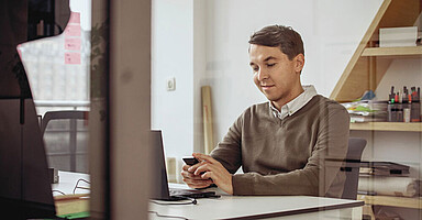 Mann im Büro. Er sitzt am Schreibtisch und schaut auf sein Smartphone.