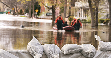 Durch Hochwasser überflutete Straße mit Sandsäcken im Vordergrund und Einsatzkräften im Wasser