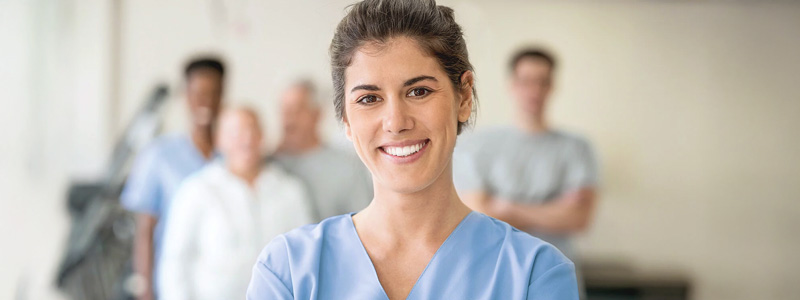 Pflegekräfte stärken: Eine Pflegekraft lächelt Betrachtende an, hinter ihr stehen weitere Mitarbeitende. Pflegekräfte stärken: Eine Pflegekraft lächelt Betrachtende an, hinter ihr stehen weitere Mitarbeitende.