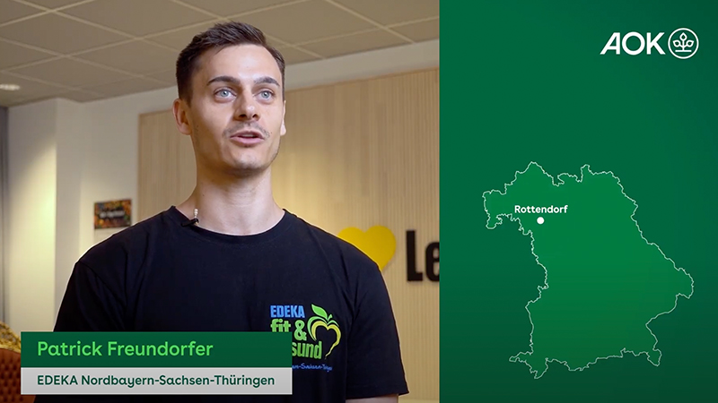 Patrick Freundorfer von EDEKA Nordbayern-Sachsen-Thüringen steht vor einer Wand mit dem EDEKA 'fit & gesund' Logo. Auf der rechten Seite des Bildes ist eine grüne Karte mit der Region Bayern und einem AOK-Logo oben rechts zu sehen.