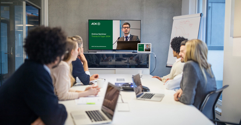 Besprechungsraum, in dem alle Teilnehmenden auf einen Monitor an der Wand schauen.