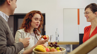 Iss mehr Vitamine Drei Personen stehen um einen Obstkorb herum und eine Frau isst eine Birne.