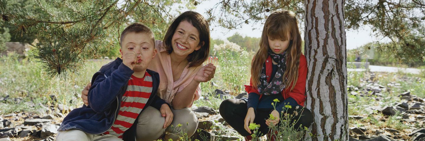 Krankenkassenwechsel lohnt sich: Lächelnde Mutter sitzt mit zwei kleinen Kindern unter einem Baum.