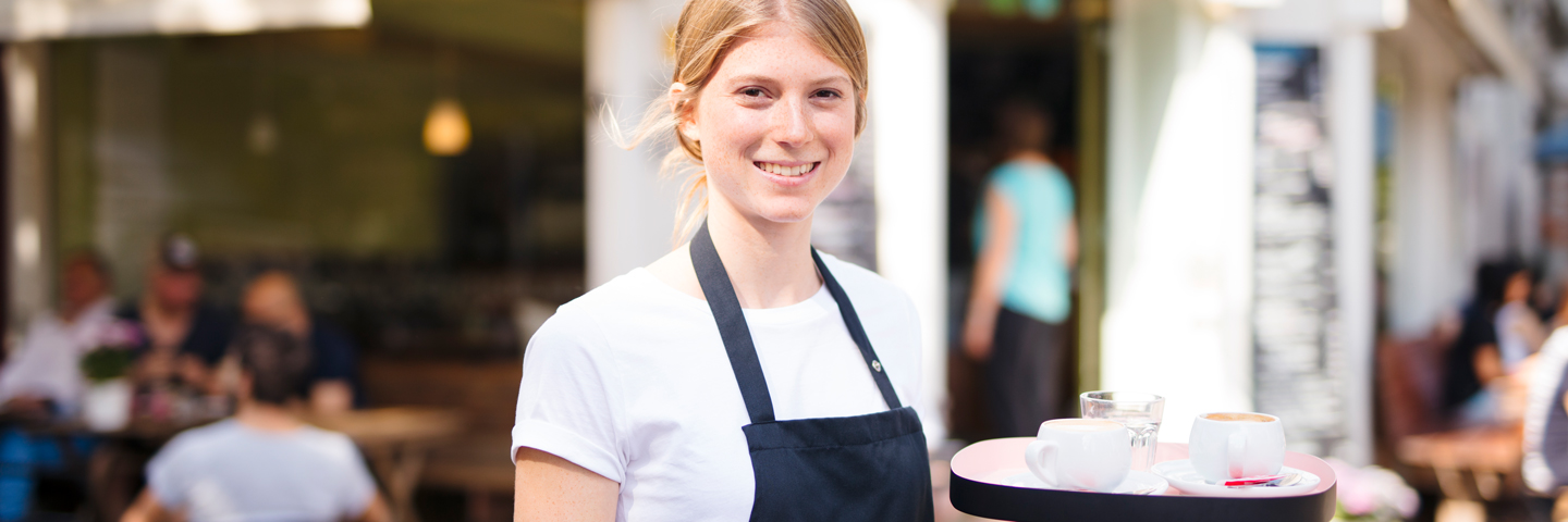 Eine junge Frau mit Schürze steht vor einem Straßencafé und trägt ein Serviertablett mit Kaffeetassen sowie eine Speisekarte.