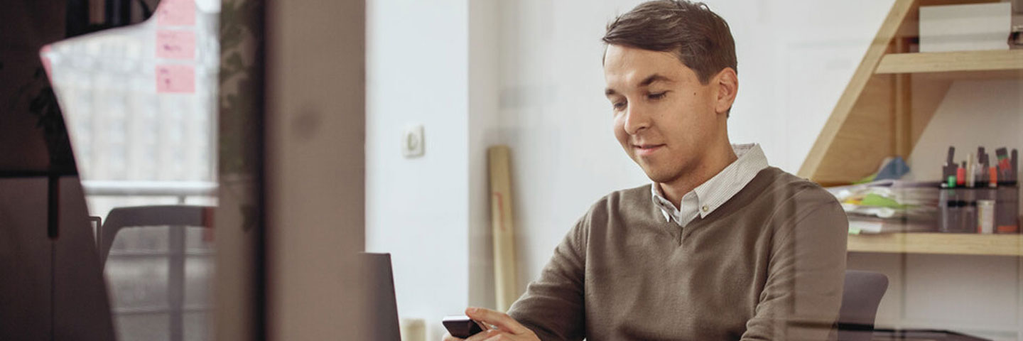 Mann im Büro. Er sitzt am Schreibtisch und schaut auf sein Smartphone.