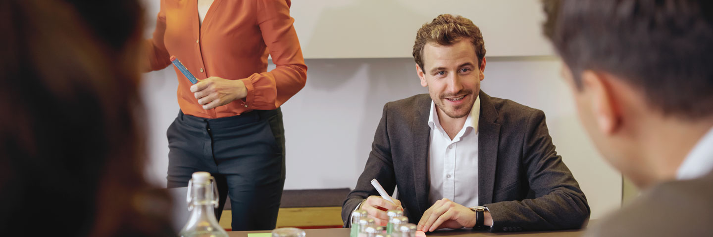 Personen am Konferenztisch. Im Fokus ein Mann, der zuversichtlich in die Runde blickt.