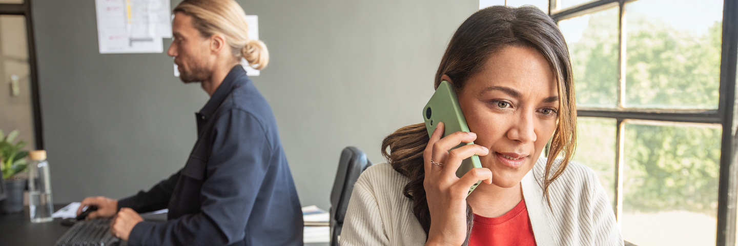 Krankenkassenwahlrecht: In einem Büro telefoniert eine Frau, ihr Kollege im Hintergrund arbeitet am PC. 