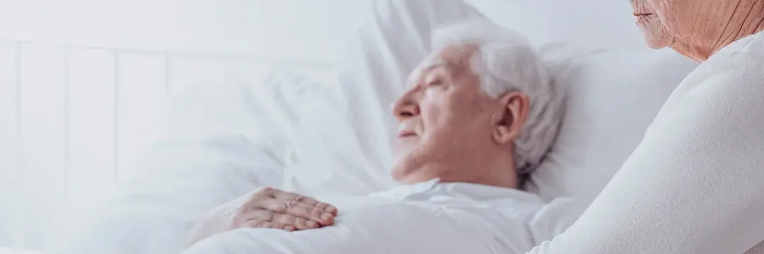 Ältere Frau unterstütz kranken Mann der im einem Krankenhausbett liegt