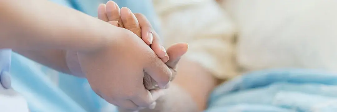 Krankenschwester sitze auf einen Pflegebett in dem eine ältere Frau liegt, beide halten ihre Hände