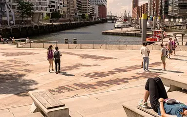 Menschen liegen und sitzen auf den Magellanterrassen in der Sommersonne und blicken auf die Elbphilharmonie und die Hafencity in Hamburg. 