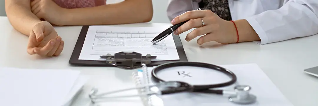 Ein Arzt und ein Patient sitzen an einem Schreibtisch und besprechen Informationen auf einem Dokument an einem Klemmbrett.
