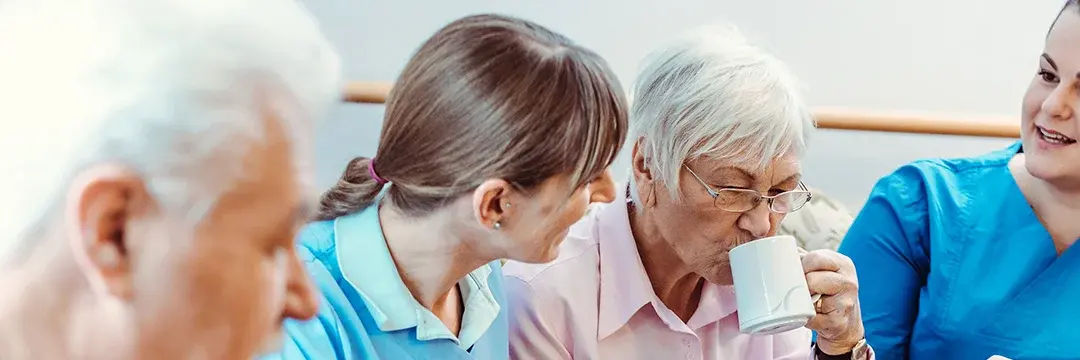 Pflegekräfte unterhalten sich mit zwei älteren Personen. Die Dame trinkt dabei aus einer Tasse Pflegekräfte unterhalten sich mit zwei älteren Personen. Die Dame trinkt dabei aus einer Tasse