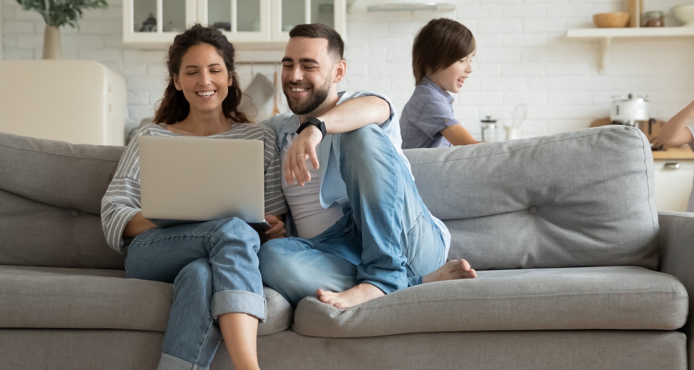 Eine junge Familie im Wohnzimmer: Die Eltern sitzen lächelnd und entspannt nebeneinander auf einem grauen Sofa und schauen auf einen Laptop, den die Frau auf ihrem Schoß hat. Um das Sofa herum spielen ihre beiden Kinder fangen, der Junge läuft dem Mädchen hinterher.