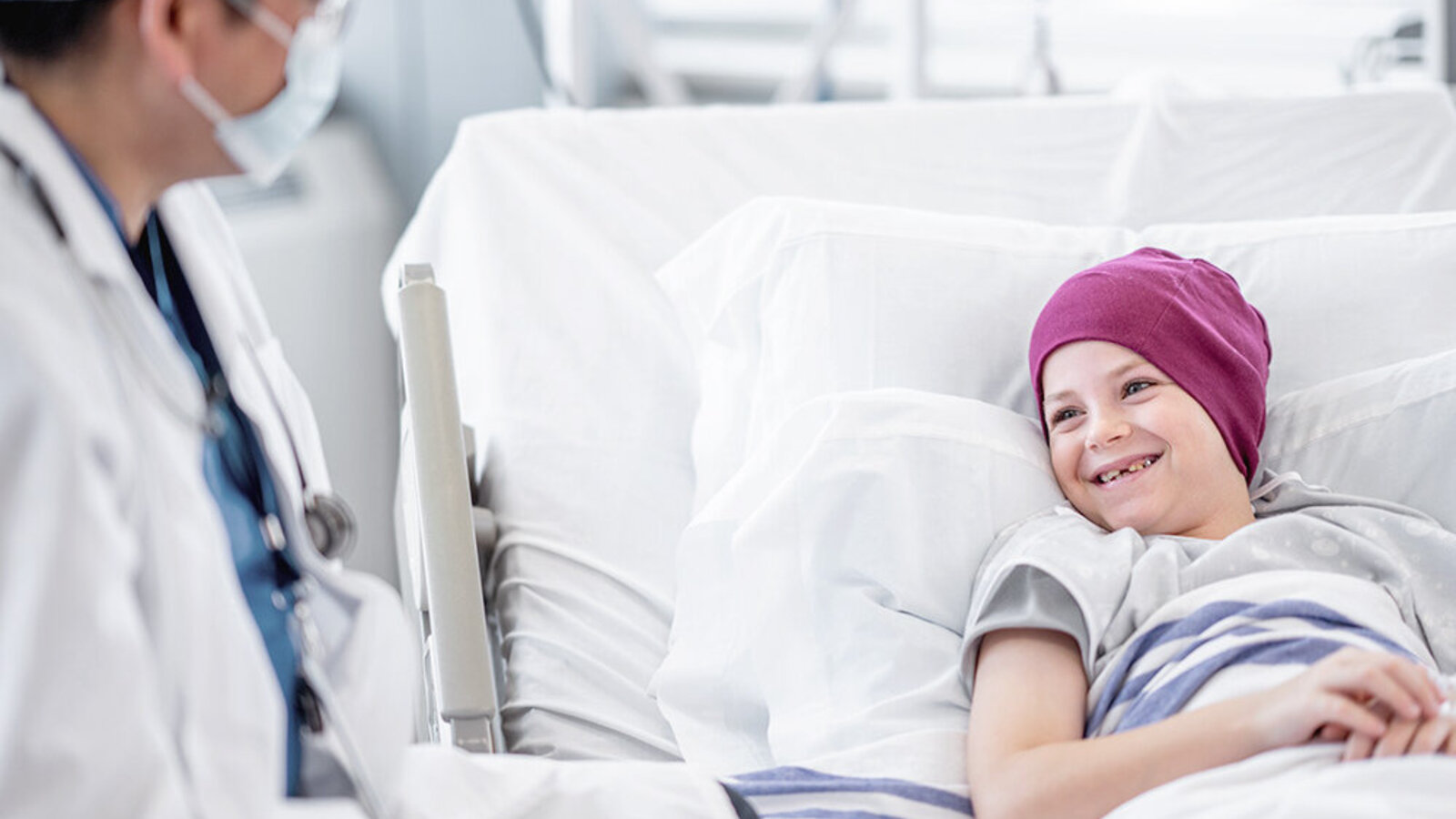 Kind mit Kopftuch im Krankenbett lacht einem Arzt zu