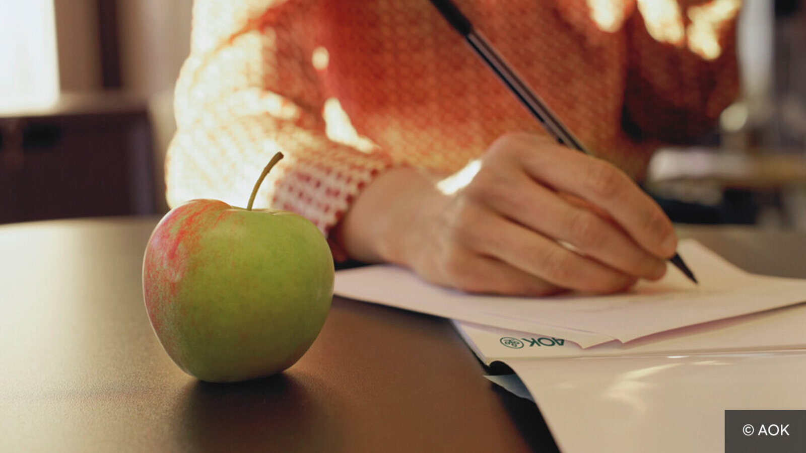 Eine Hand hält einen Bleistift und notiert etwas auf einem AOK-Schreibblock. 