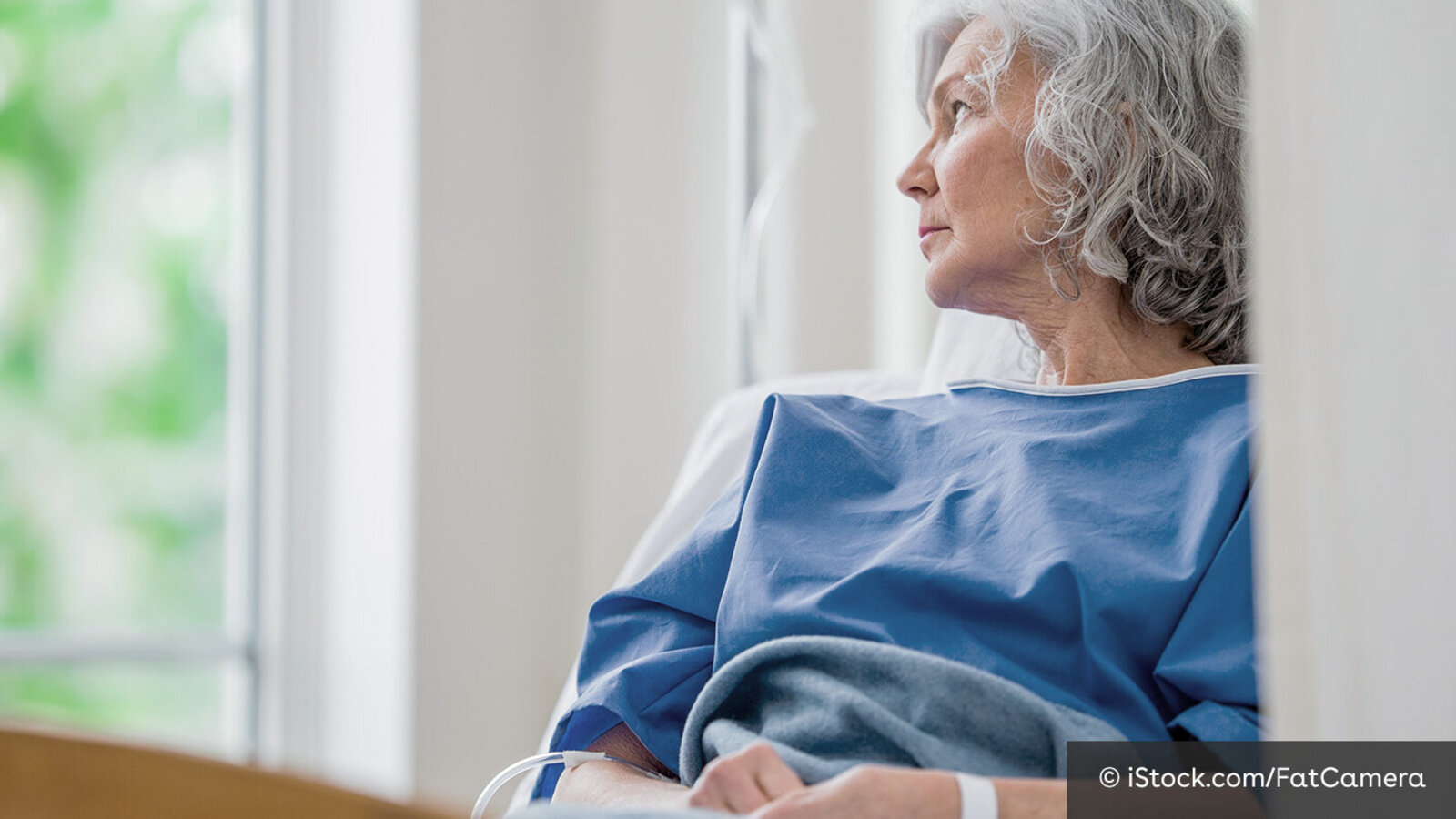 Eine Frau mit weißgrauen Haaren liegt in Krankenhauskleidung im Klinikbett und schaut aus dem Fenster. Neben ihr steht ein Tropf, in ihrem Arm steckt eine Kanüle. 