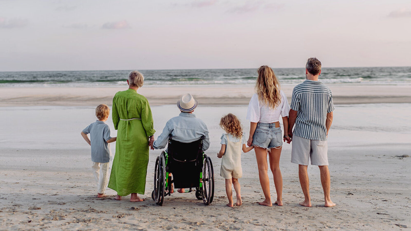Familie steht am Strand und schaut aufs Meer, sie halten sich an den Händen, der Großvater sitzt im Rollstuhl