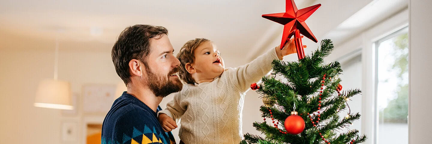 Ein Vater steht in einem Wohnzimmer und hält sein kleines Kind auf dem Arm, das einen roten Stern auf die Spitze eines Weihnachtsbaumes steckt.