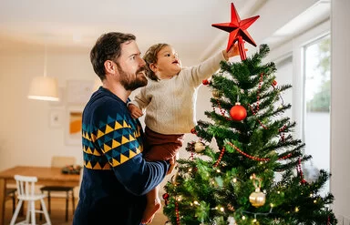 Ein Vater steht in einem Wohnzimmer und hält sein kleines Kind auf dem Arm, das einen roten Stern auf die Spitze eines Weihnachtsbaumes steckt.