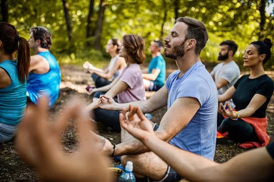 Acht junge Männer und Frauen sitzen im Schneidersitz in der Yoga-Pose Sukhasana in einem grünen Park. Sie tragen helle, kurzärmlige Sportkleidung, haben entspannte Gesichter und die Augen geschlossen.
