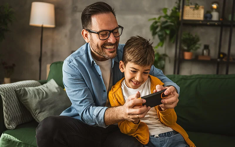 Ein Vater sitzt mit seinem Sohn auf dem Sofa. Der Vater umarmt das Kind von hinten und beide halten mit ihren Händen ein Smartphone. Beide schauen amüsiert auf den Bildschirm.