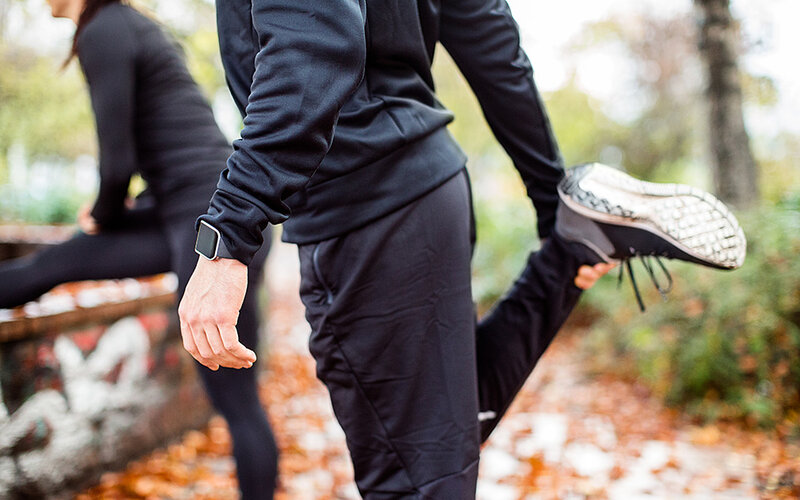 Ein Sportler in dunkler Sportkleidung im Park zieht seinen rechten Fuß zur Dehnung an den Po. Im Hintergrund ist verschwommen eine Sportlerin zu sehen, die ihr Bein zur Dehnung ausgestreckt hat.