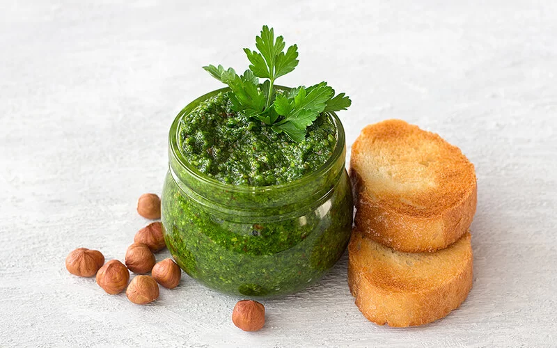 Auf einem marmorierten Untergrund steht ein Glas mit grünem Pesto, garniert mit frischer Petersilie. Daneben liegen einige ganze Haselnüsse und zwei getoastete Baguettescheiben.