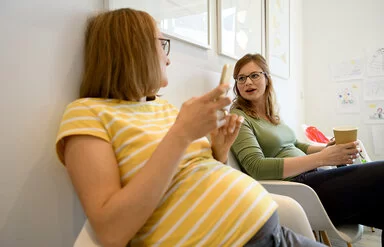 Zwei Frauen sitzen im Wartezimmer und unterhalten sich; die eine hält ein Smartphone in der rechten Hand.