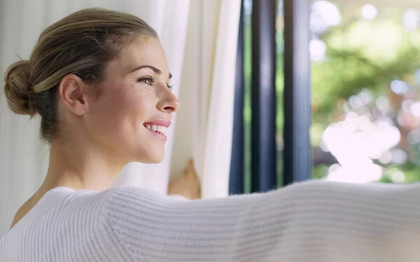 Eine Frau steht am Fenster, während sie lächelnd die Vorhänge aufzieht und nach draußen blickt.