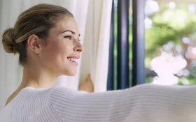 Eine Frau steht am Fenster, während sie lächelnd die Vorhänge aufzieht und nach draußen blickt.