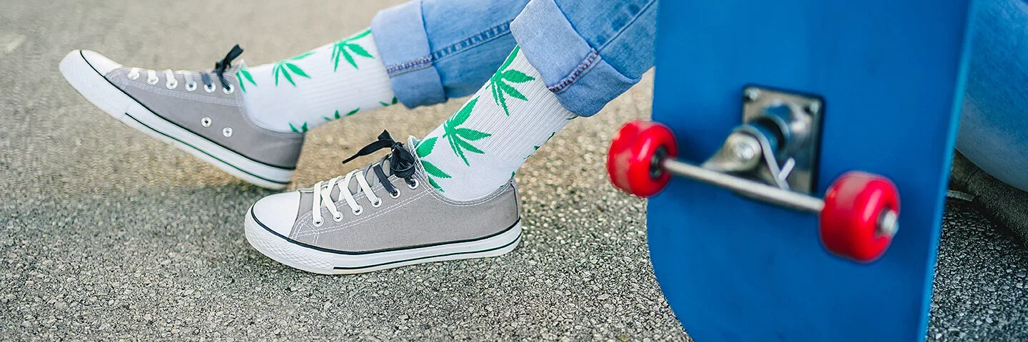 Im Bildausschnitt sind die Beine eines Menschen zu sehen. Er trägt Blue Jeans und graue Sneaker. Auf den weißen Socken sind grüne Marihuana-Pflanzen zu sehen. Er sitzt auf einer Straße und hat die Beine angewinkelt. Daneben ist ein blaues Skateboard mit roten Rädern zu sehen. 