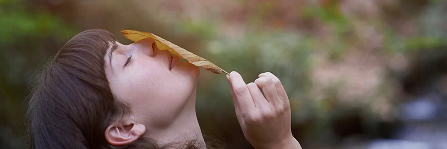 Frau hebt Kopf nach oben, riecht an Blatt in ihrer Hand, verschwommener Wald-Hintergrund.