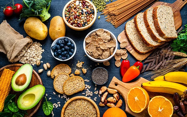 Verschiedene nährstoffreiche Lebensmittel auf einer schwarzen Schieferplatte: zwei Brotsorten, getrocknete Hülsenfrüchte, Getreide, Heidelbeeren, Chiasamen, Dinkelflakes, Avocado, Maiskolben, Kartoffeln, Orangen, Bananen, Vollkornnudeln, Brokkoli, Tomaten, Mini-Paprika, Paranüsse, Sonnenblumenkerne und Kürbiskerne.