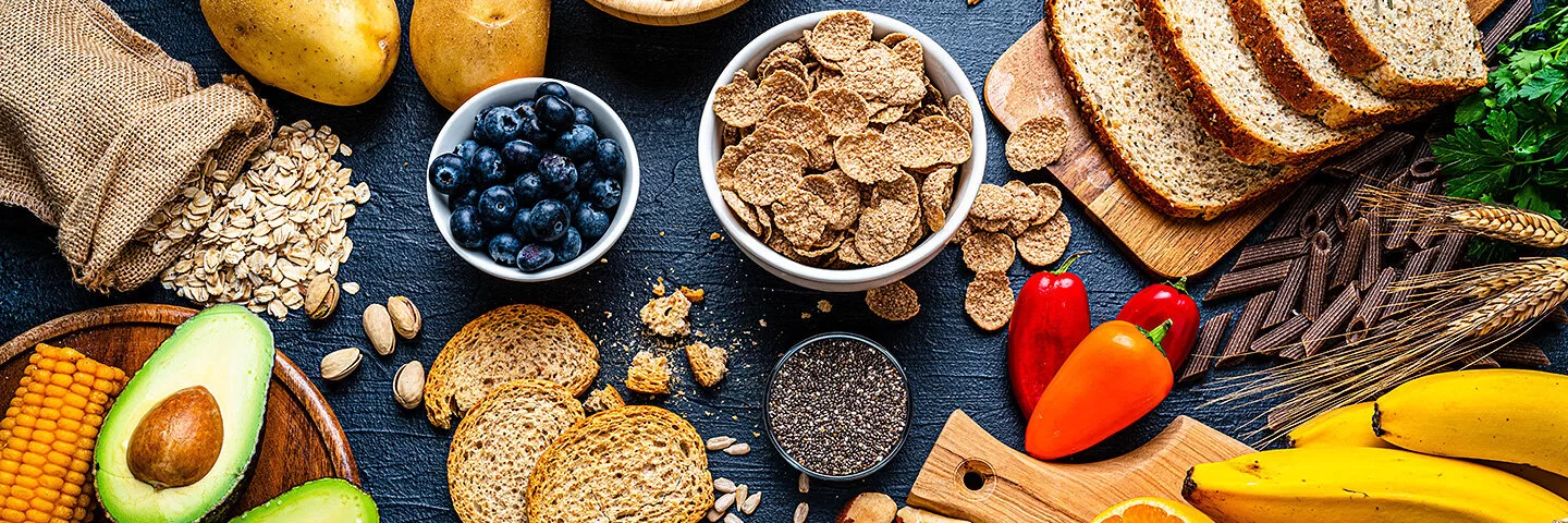 Verschiedene nährstoffreiche Lebensmittel auf einer schwarzen Schieferplatte: zwei Brotsorten, getrocknete Hülsenfrüchte, Getreide, Heidelbeeren, Chiasamen, Dinkelflakes, Avocado, Maiskolben, Kartoffeln, Orangen, Bananen, Vollkornnudeln, Brokkoli, Tomaten, Mini-Paprika, Paranüsse, Sonnenblumenkerne und Kürbiskerne.