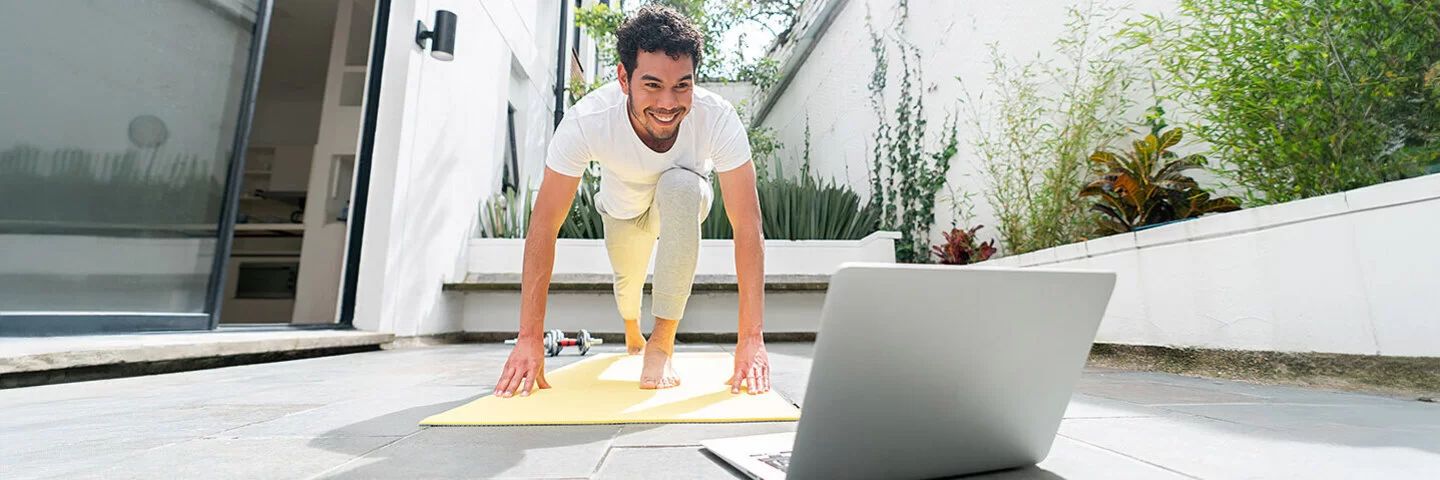 Junger Mann, der zu Hause vor seinem Laptop auf der Terasse nach einem Online-Fitnesskurs trainiert