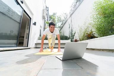 Junger Mann, der zu Hause vor seinem Laptop auf der Terasse nach einem Online-Fitnesskurs trainiert