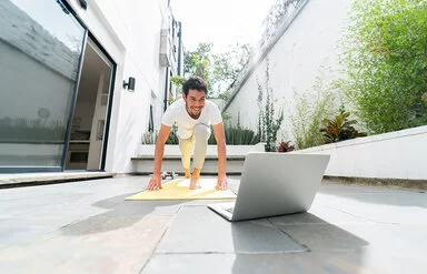 Junger Mann, der zu Hause vor seinem Laptop auf der Terasse nach einem Online-Fitnesskurs trainiert