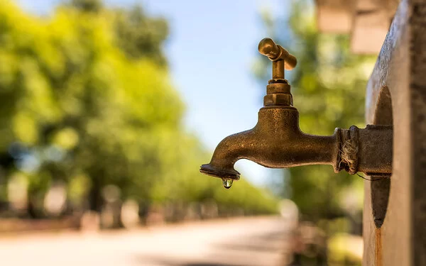 Abbildung eines öffentlich zugänglichen Wasserhahns, im Hintergrund befinden sich ein Weg und Bäume.