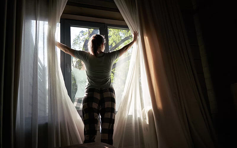 Eine Frau im Pyjama zieht die Vorhänge ihres Schlafzimmers beiseite und lässt das helle Morgenlicht herein.