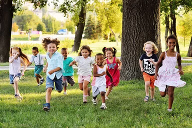 Neun Kinder im Kindergarten und Grundschulalter rennen mit fliegenden Haaren um die Wette – sie laufen auf die Kamera zu. Sie sind sommerlich in bunten T-Shirts, Shirts und Kleidern gekleidet.