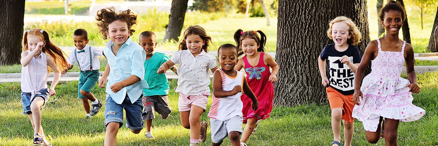 Neun Kinder im Kindergarten und Grundschulalter rennen mit fliegenden Haaren um die Wette – sie laufen auf die Kamera zu. Sie sind sommerlich in bunten T-Shirts, Shirts und Kleidern gekleidet.